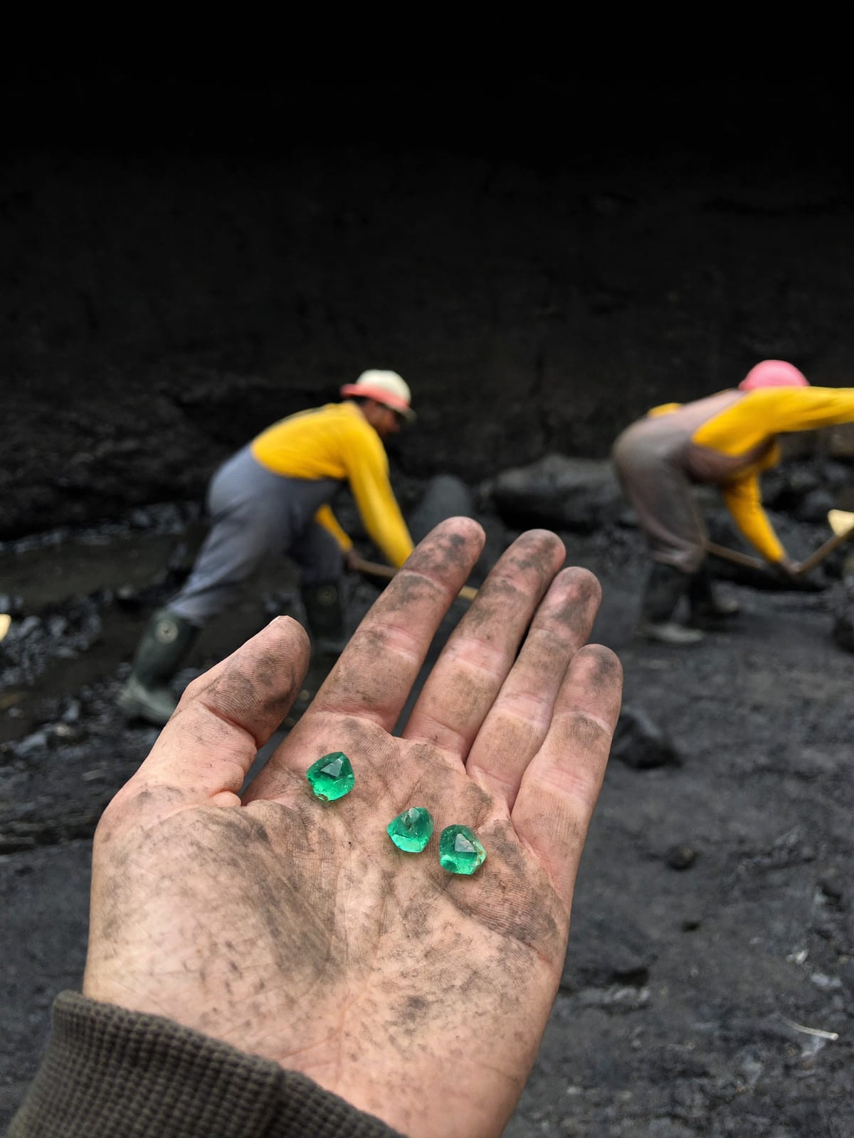 Mano de un minero mostrando tres esmeraldas talladas, con mineros trabajando en la mina al fondo