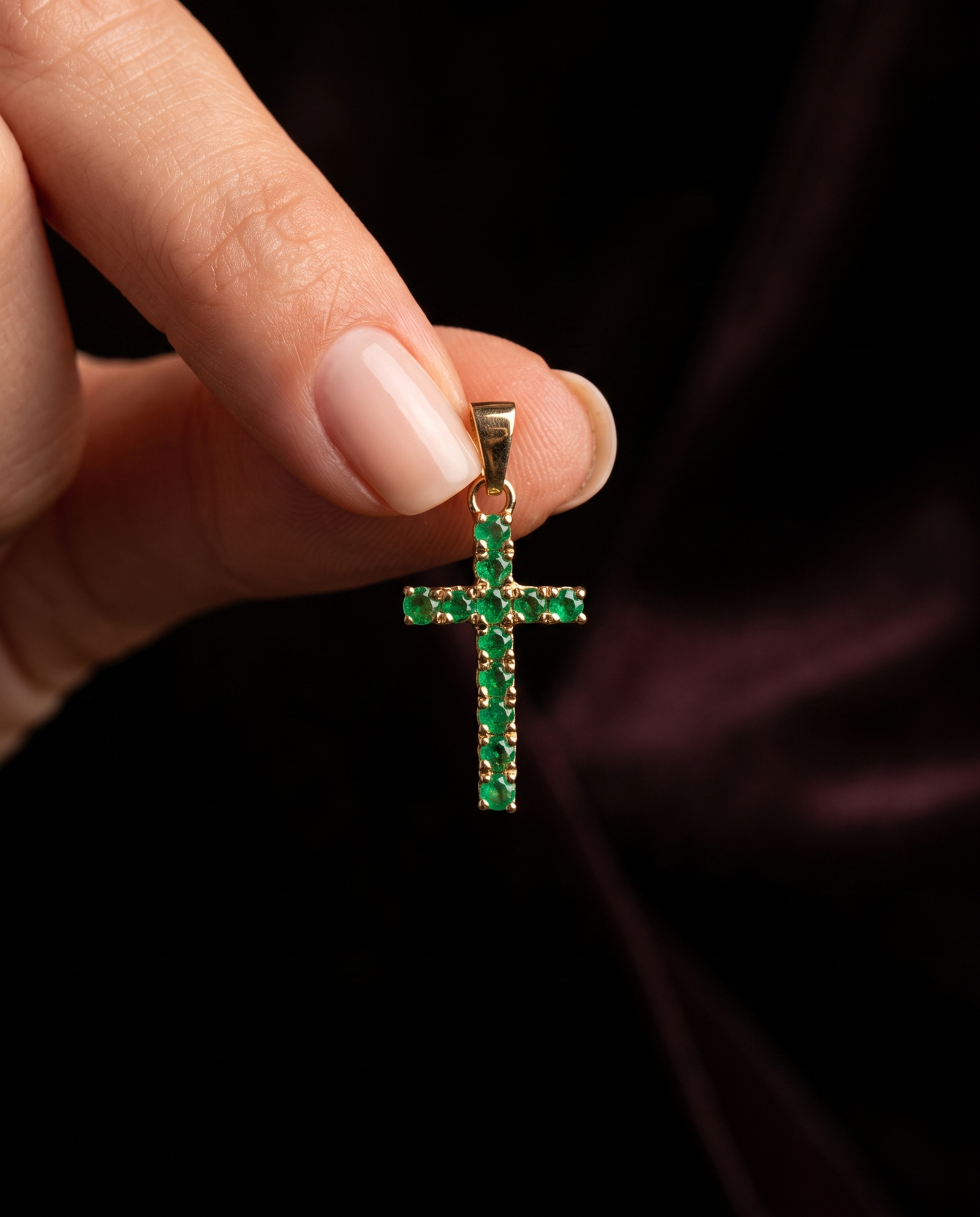 Dije cruz de esmeraldas sostenido entre los dedos sobre fondo oscuro
